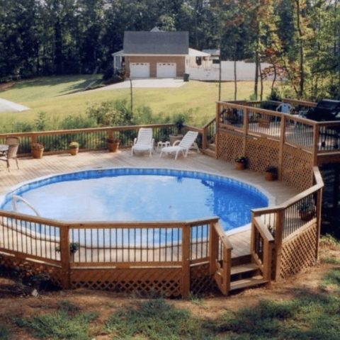 A round above-ground pool is surrounded by a wooden deck with stairs, lounge chairs, potted plants, a table with an umbrella, and a railing. The deck is set in a grassy, wooded backyard with a house in the background.